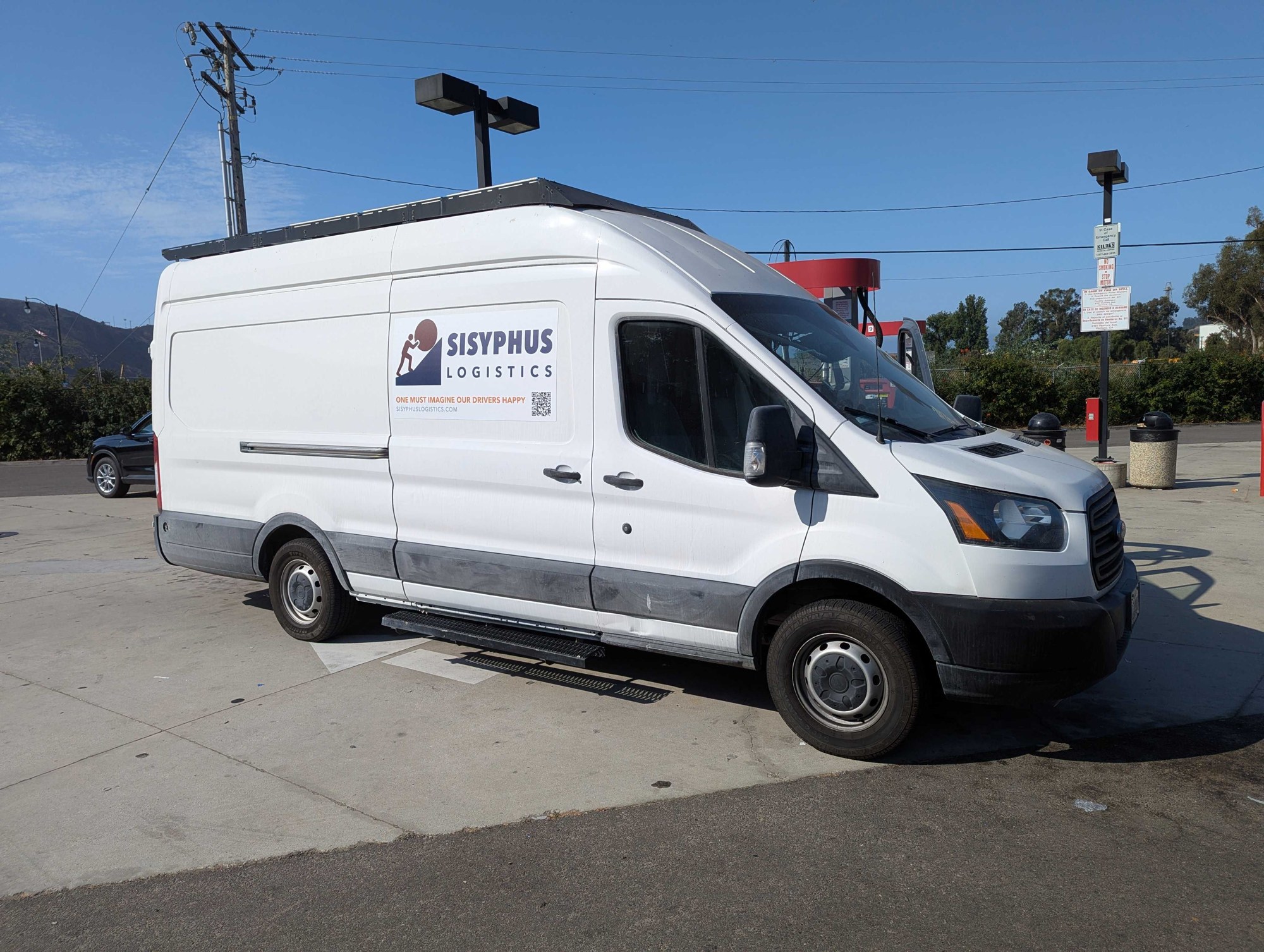 Unit 001, a white high-roof van wearing the Sisyphus Logistics door magnet, parked at a fueling stop.